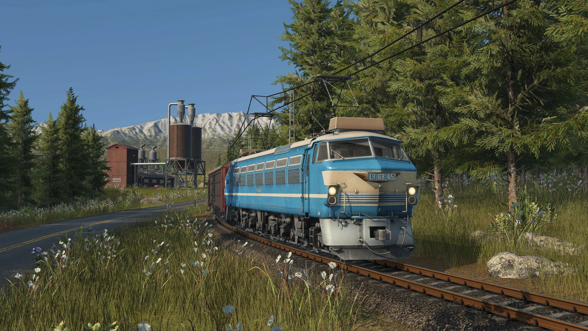 A blue electric freight locomotive passing an industrial facility in a forested mountain landscape